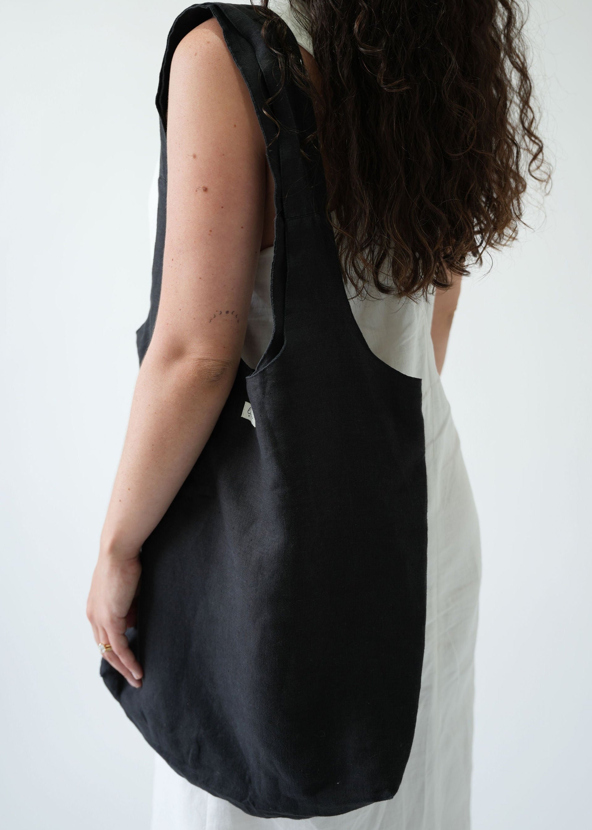 A woman wearing a white sleeveless dress carries a large black canvas shoulder tote bag against a plain white background.
