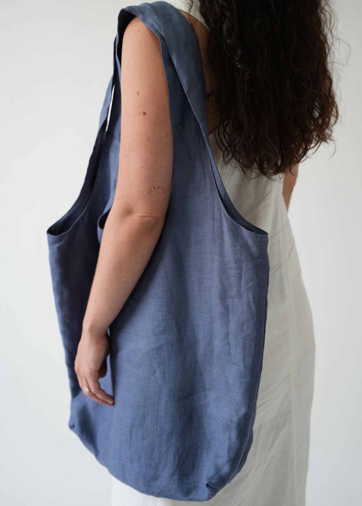 A woman shown from the back wearing a large, slouchy blue linen tote bag over her shoulder, paired with a white dress, against a plain light gray background.