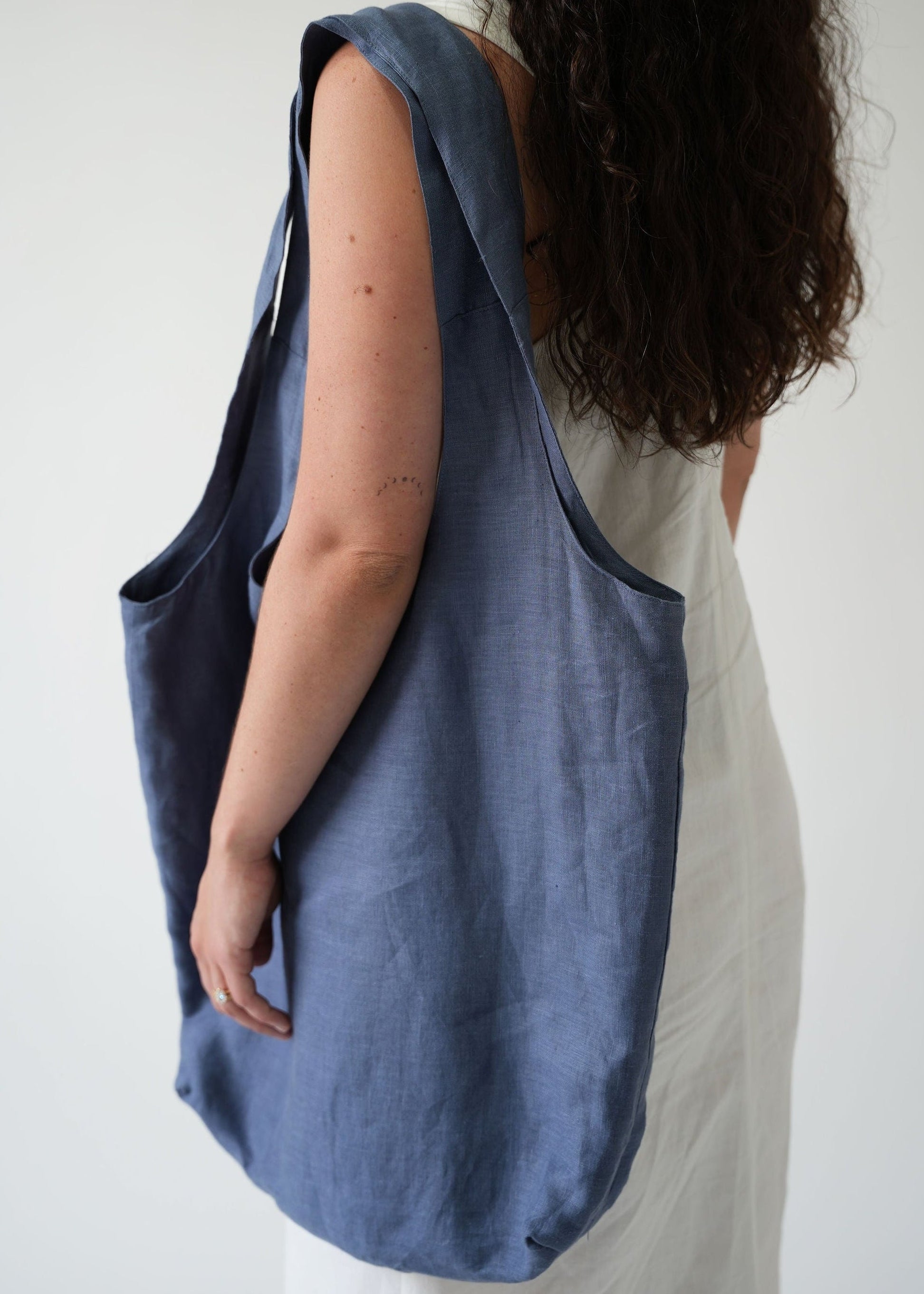 A woman shown from the back wearing a large, slouchy blue linen tote bag over her shoulder, paired with a white dress, against a plain light gray background.