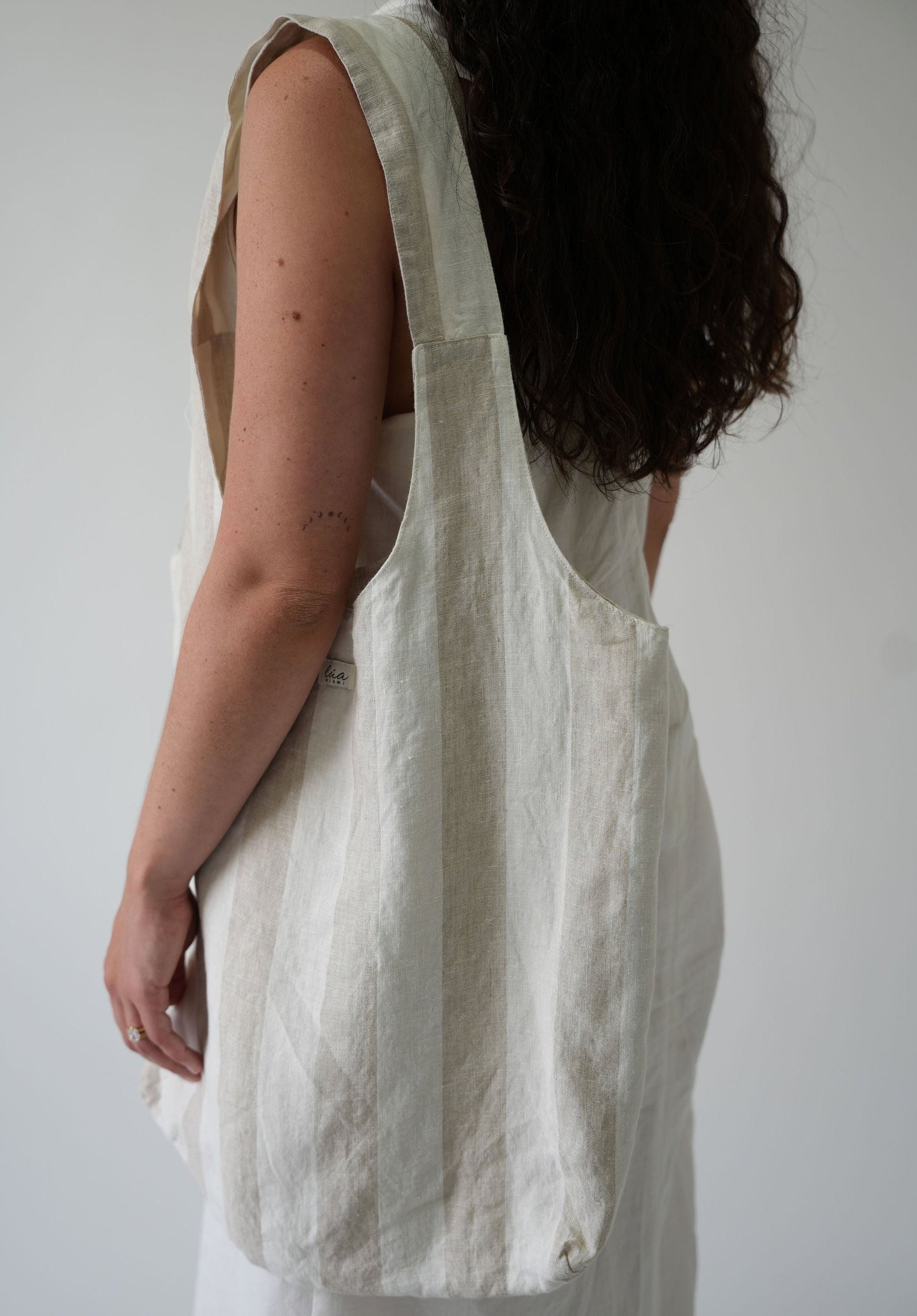 A woman shown from the back wearing a large, off-white linen shoulder bag over a white dress against a plain light gray background.