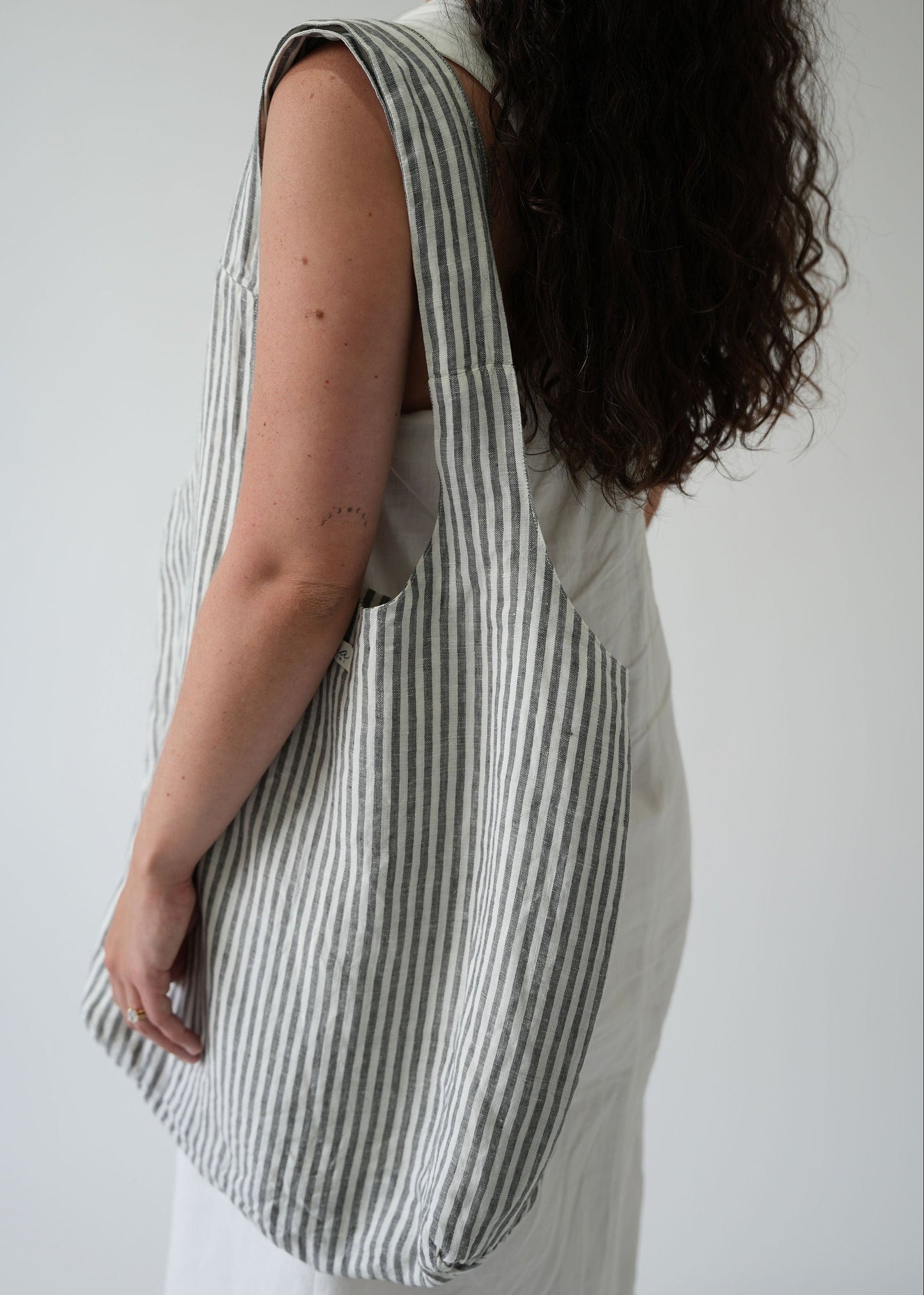 A woman shown from the back wearing a large gray and white striped fabric tote bag over her shoulder, against a plain light gray background.