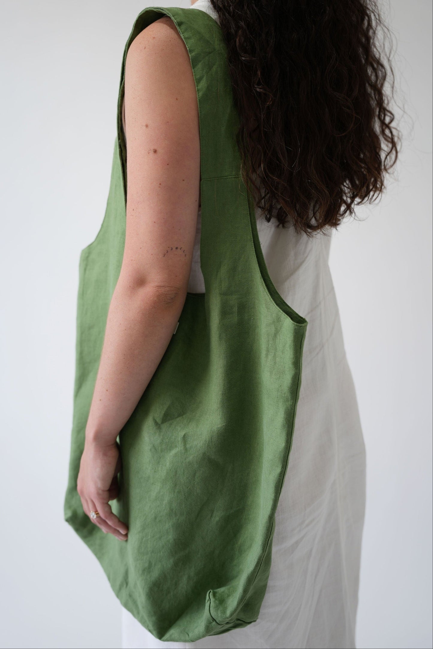 A woman wearing a white dress is shown from the side/back, carrying a large olive green linen shoulder bag over her shoulder against a plain light gray background.