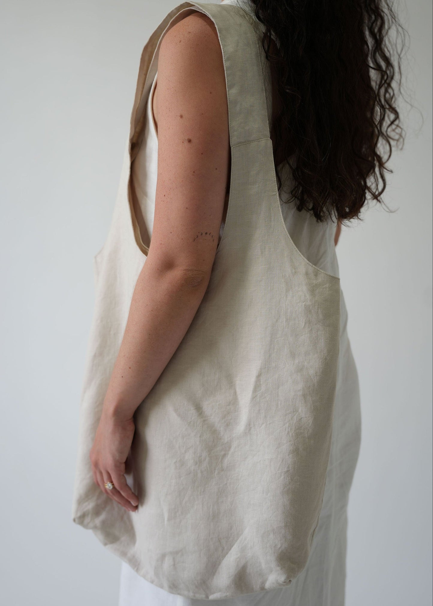 A woman shown from the side and back wearing a large, unstructured linen tote bag in a natural off-white color. The bag has a wide shoulder strap and is styled over a white top and white trousers against a plain light gray background.
