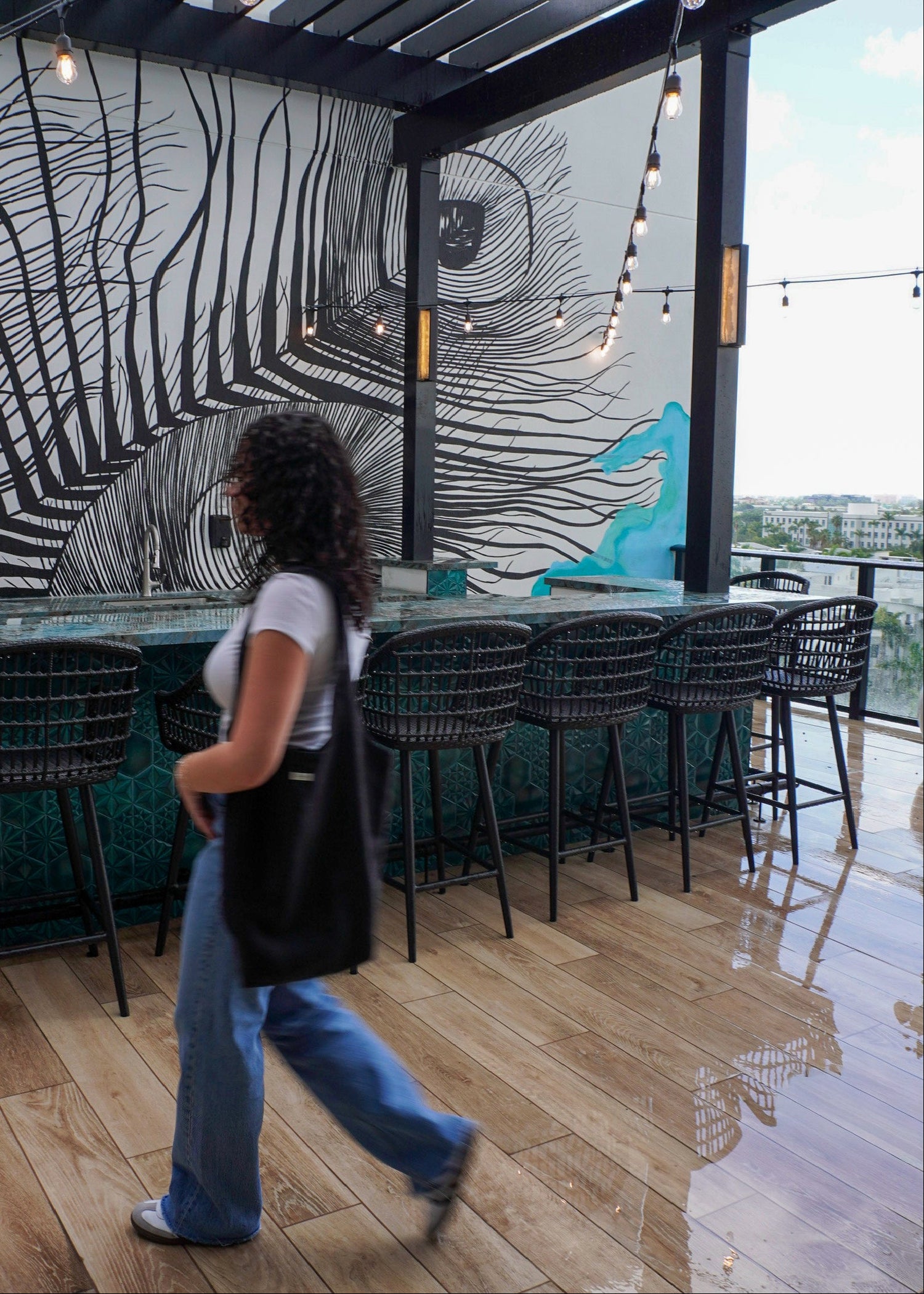 A woman walks past a modern rooftop bar with black metal pergola beams, hanging string lights, black woven bar stools, and a large black-and-white feather mural on the back wall.