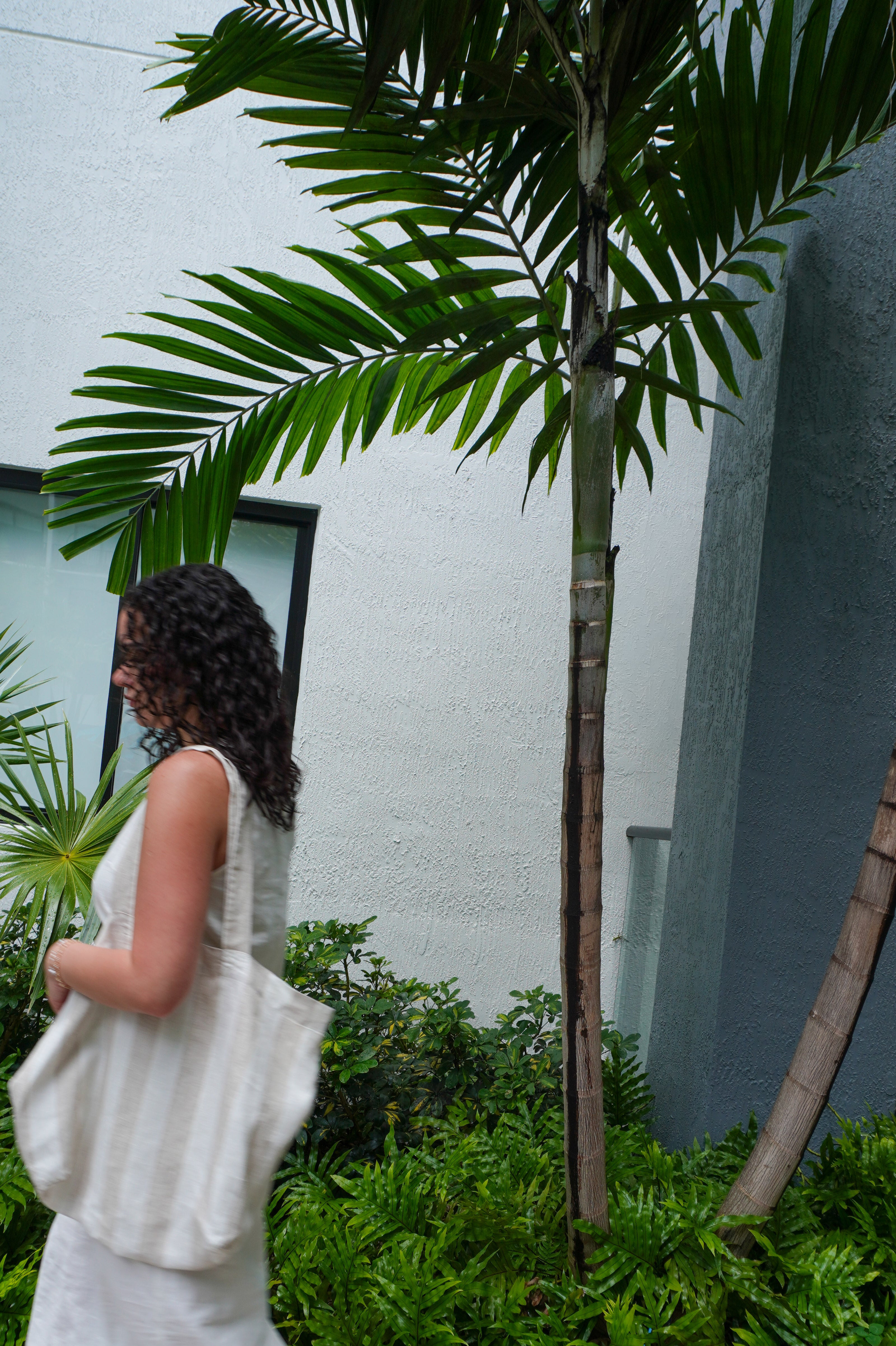 A woman in a white sleeveless dress walks past a tall palm tree in a garden setting, carrying a large white tote bag.