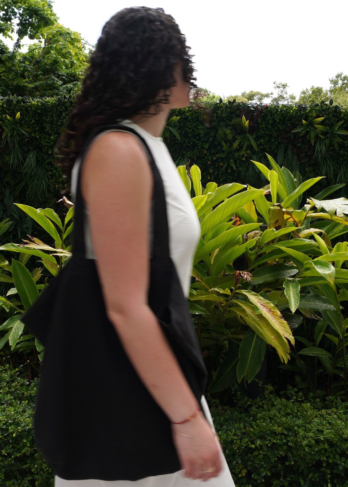 A woman in a white sleeveless dress is shown from the side, carrying a large black tote bag over her shoulder in an outdoor garden setting with green foliage.