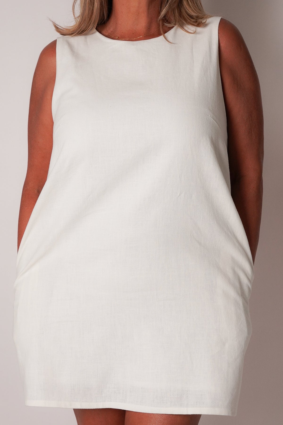A sleeveless white linen dress with side pockets, shown on a model against a neutral studio background.