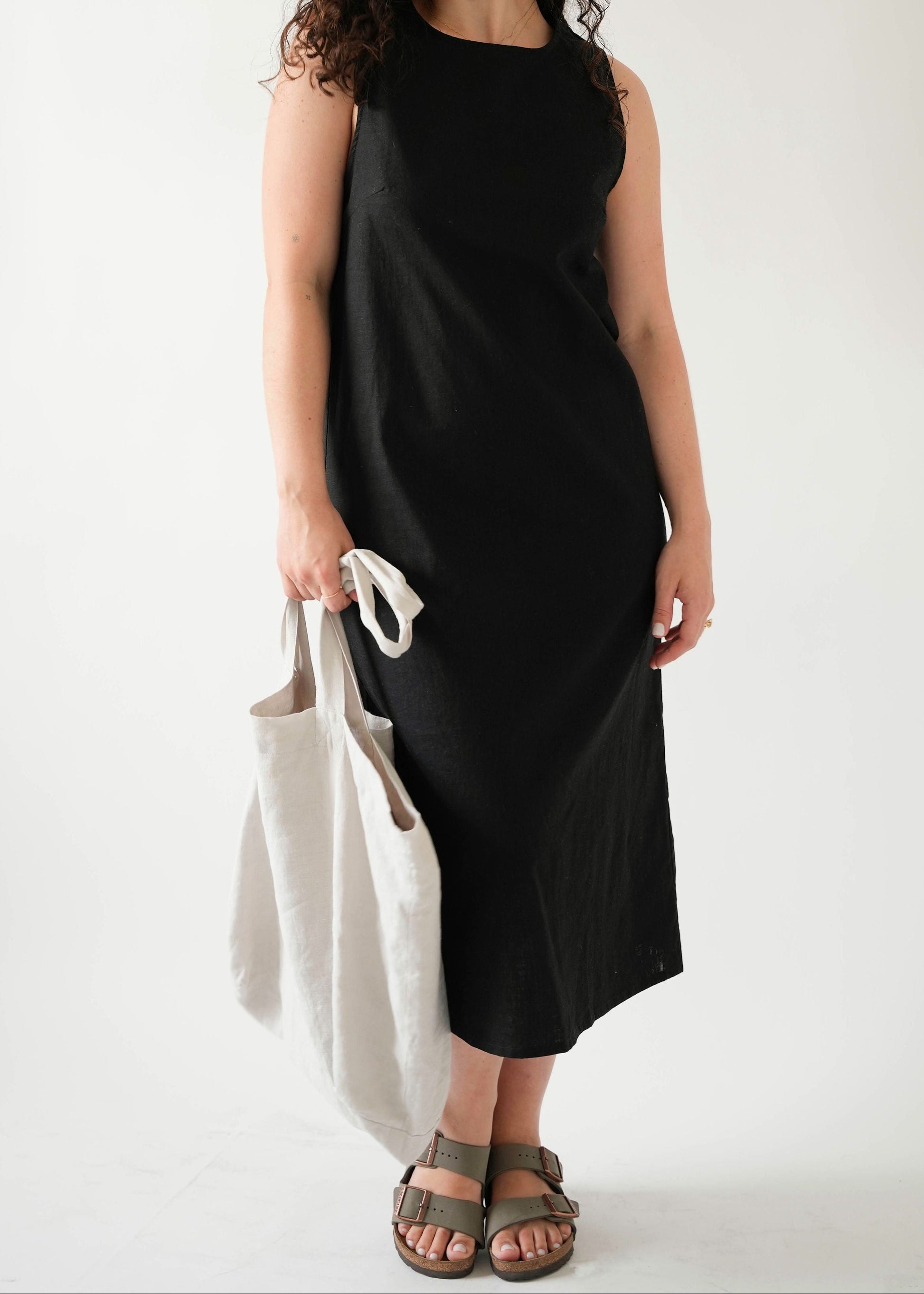 Woman wearing black linen midi dress, crew neck, sleeveless, holding natural linen tote bag, white studio background — Lúa Miami