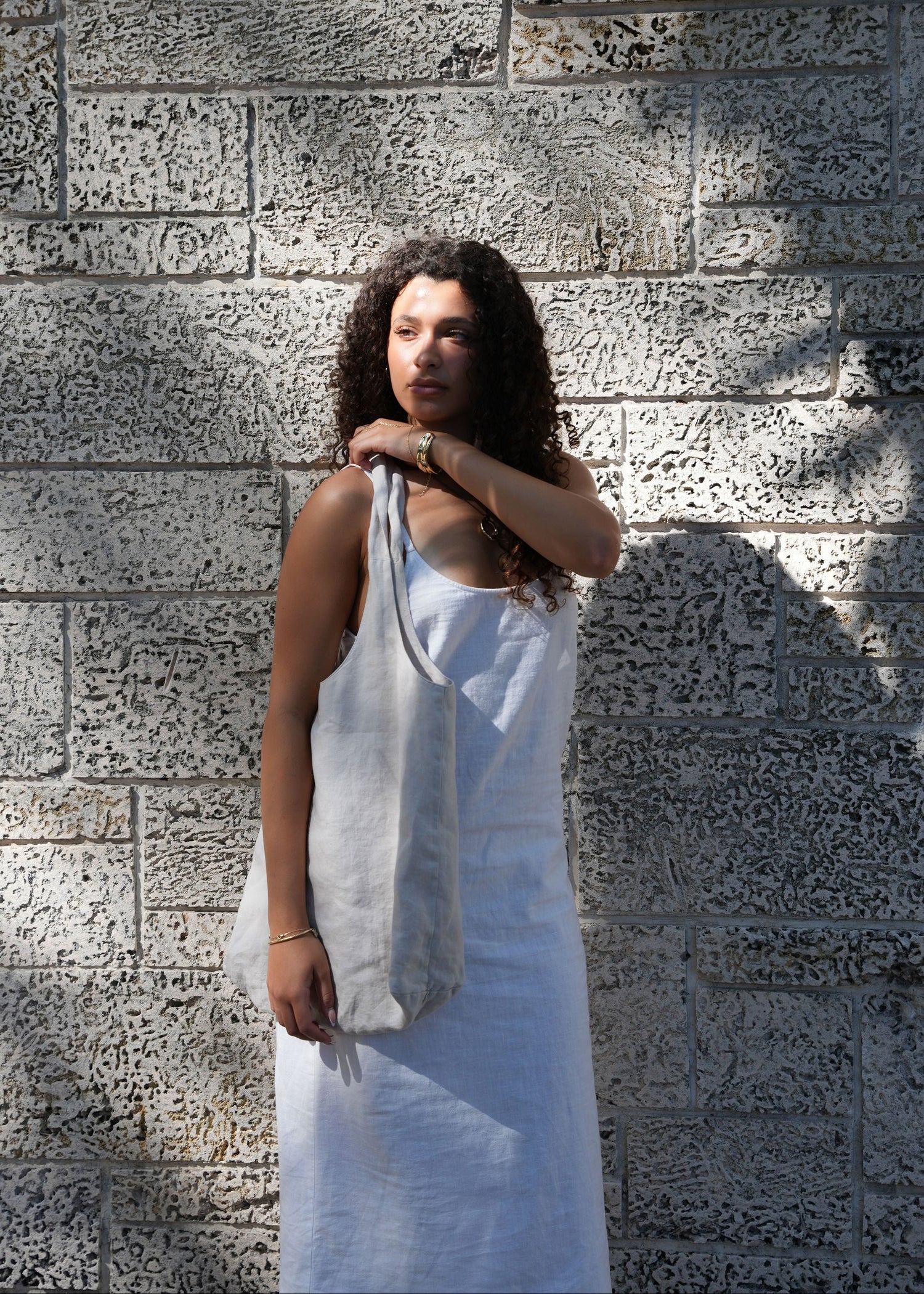A woman with curly dark hair stands against a textured stone wall, wearing a white sleeveless dress and carrying a large off-white tote bag over her shoulder.