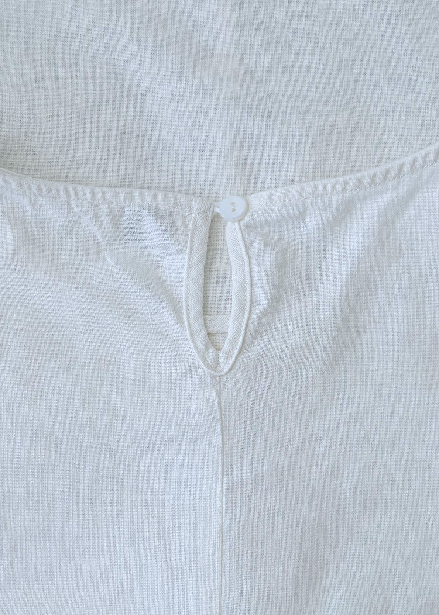 A close-up of a white sleeveless top or dress laid flat, showing the back neckline with a small button closure and stitched edge details.