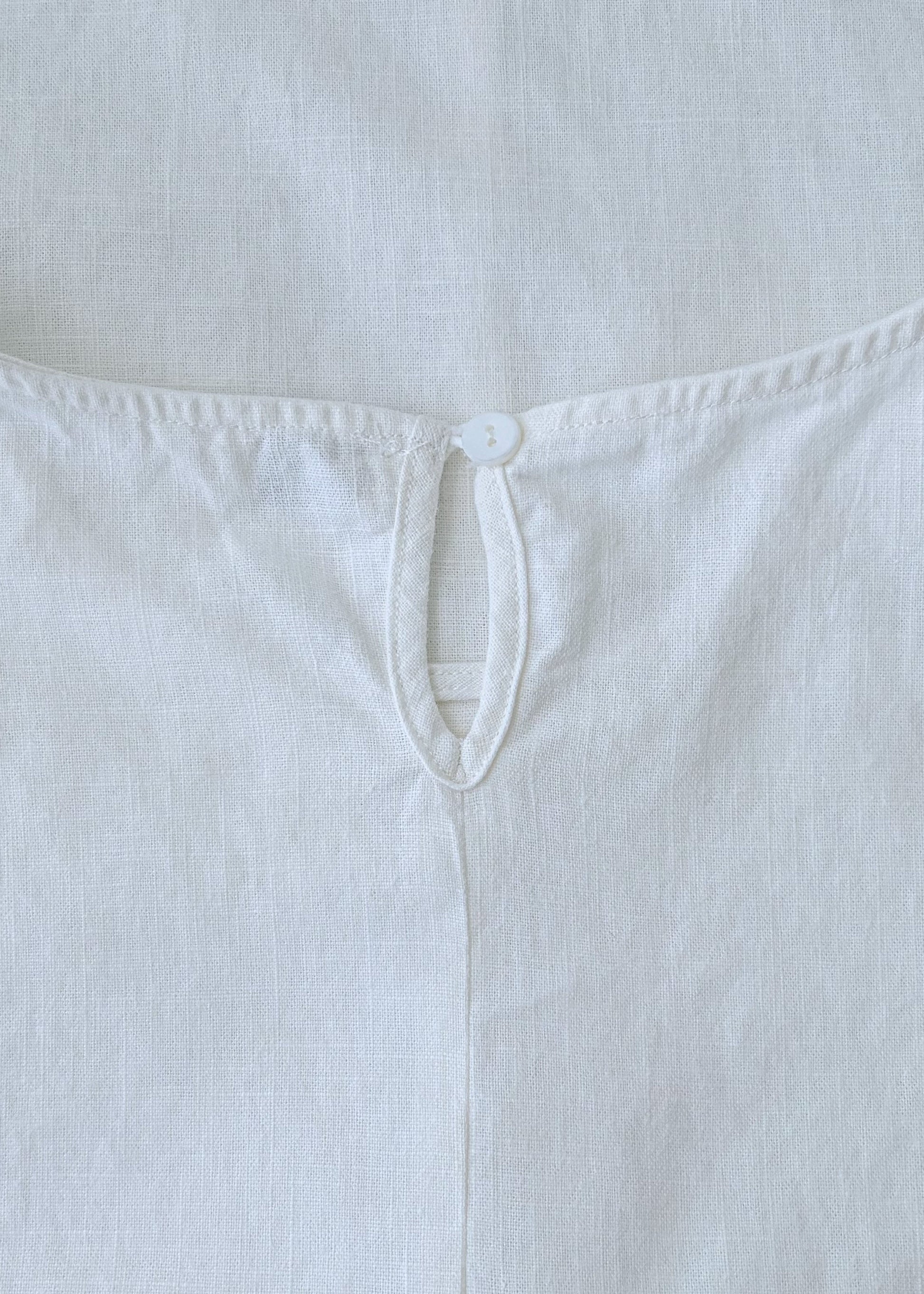 A close-up of a white sleeveless top or dress laid flat, showing the back neckline with a small button closure and stitched edge details.