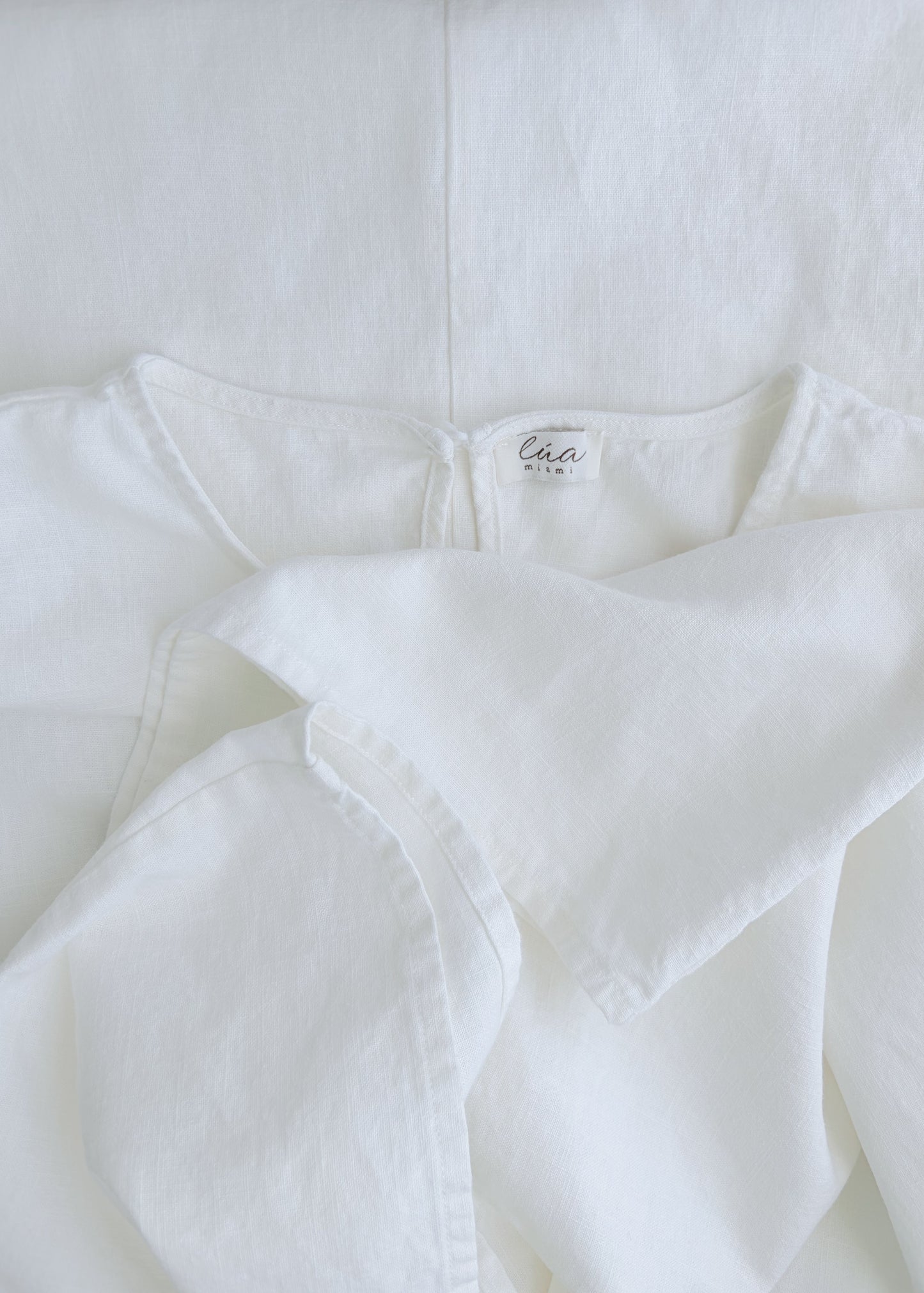 A close-up of a white cotton garment, showing the inner neck label. The fabric is softly draped, highlighting its textured, lightweight weave.