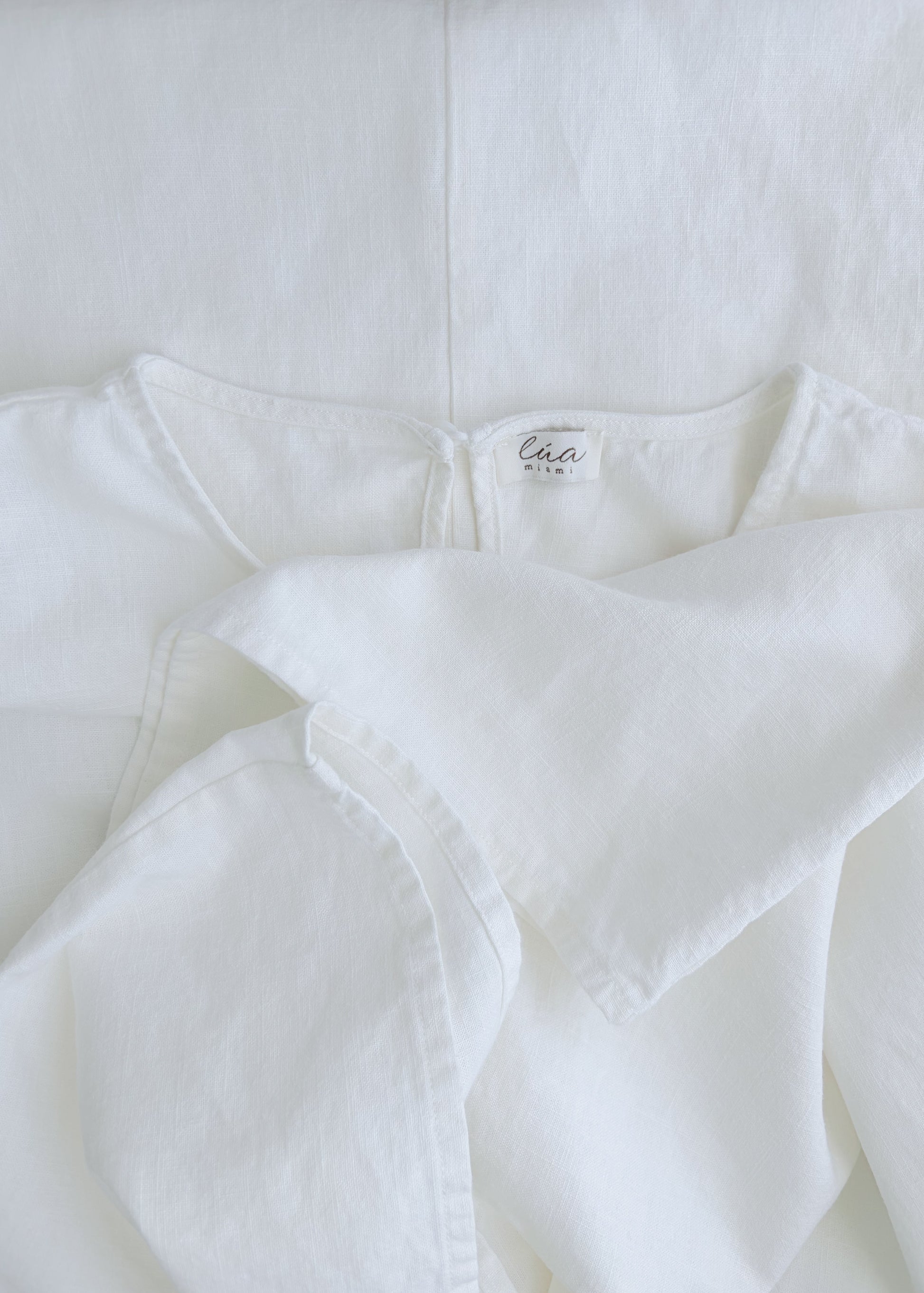 A close-up of a white cotton garment, showing the inner neck label. The fabric is softly draped, highlighting its textured, lightweight weave.