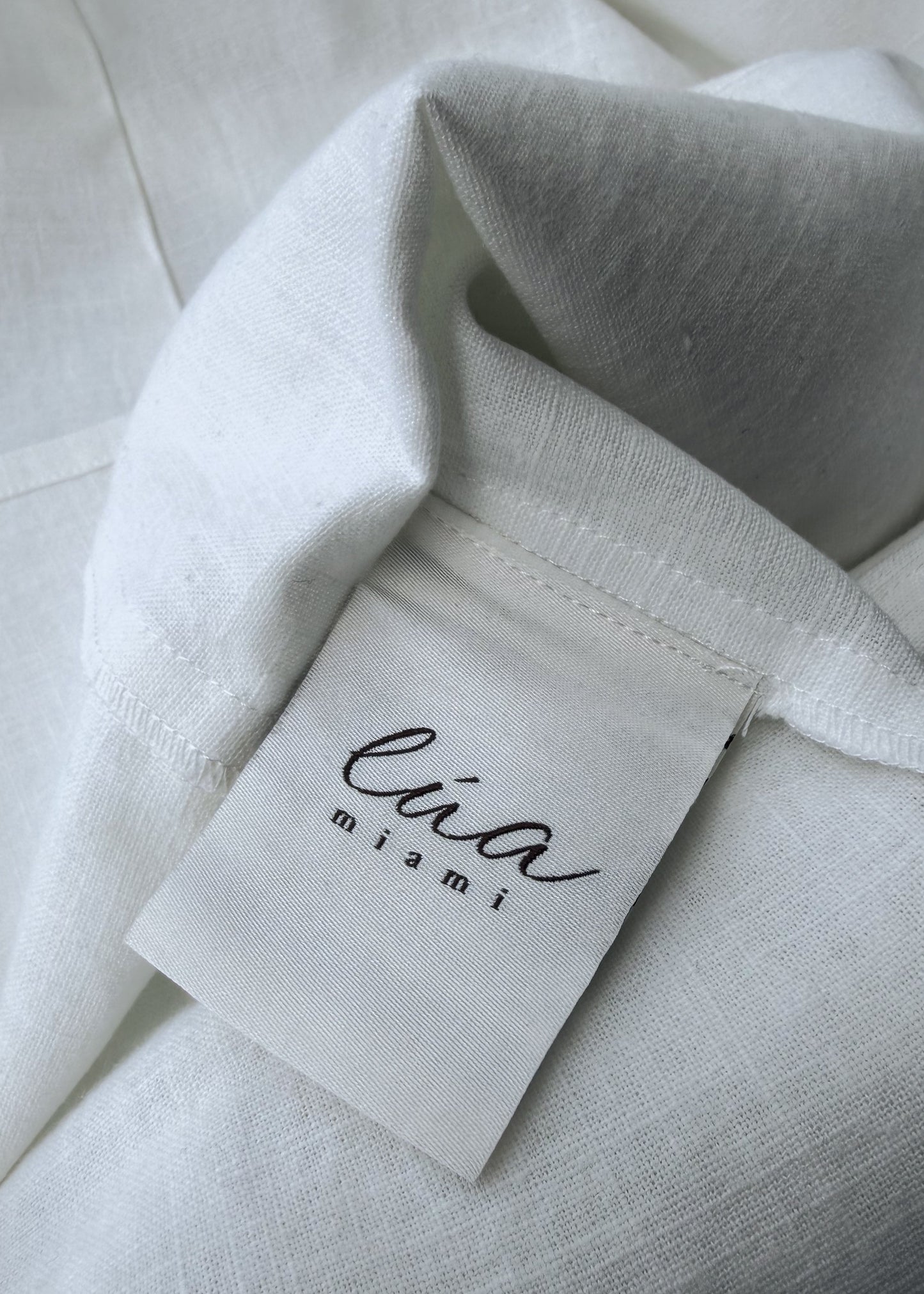 Close-up of a white textured fabric garment with a sewn-on fabric label reading 'lua miami' in black script.