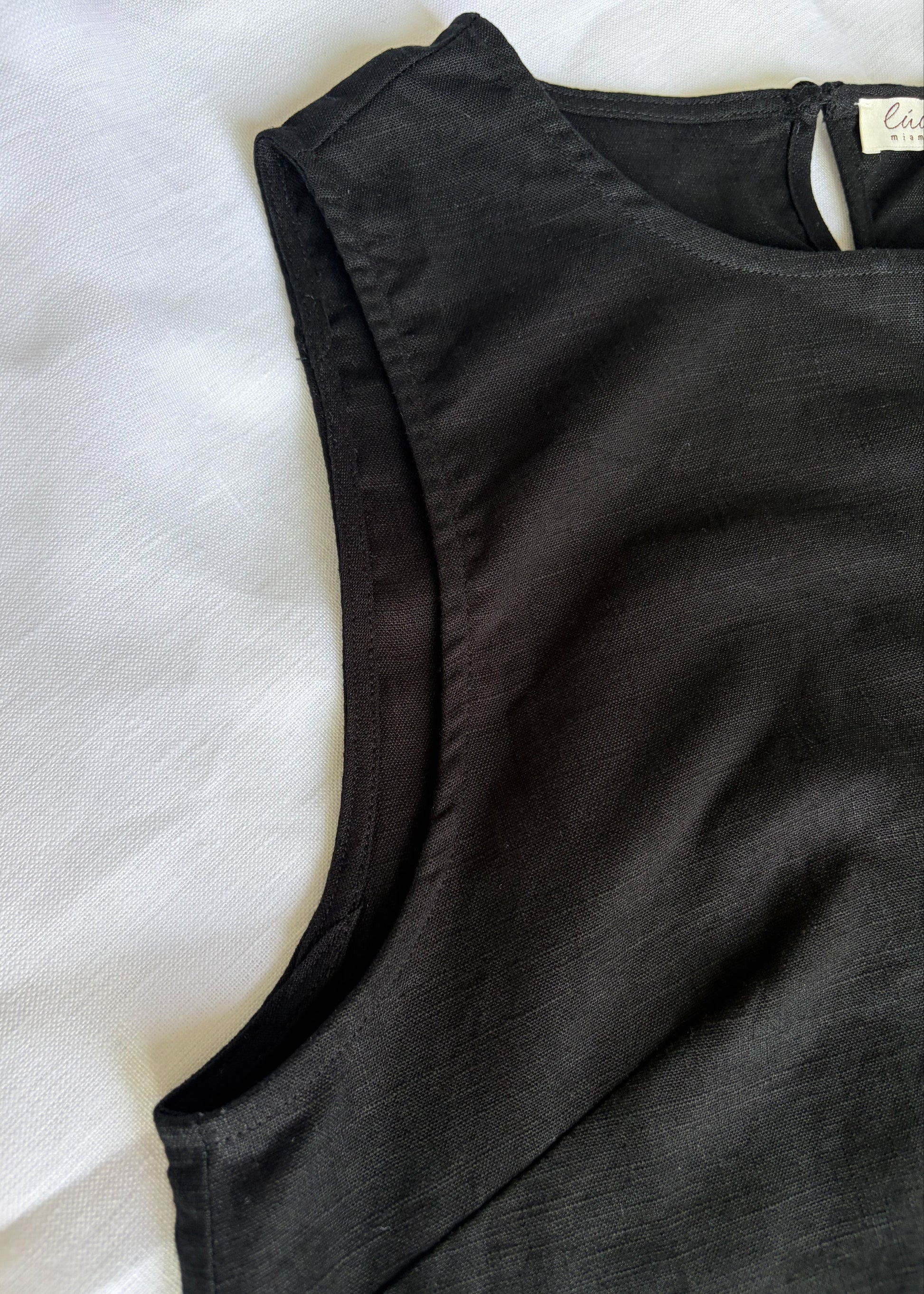 A close-up of a black sleeveless linen dress laid flat on a white fabric background, showing the shoulder seam, neckline, and a small interior neck label.
