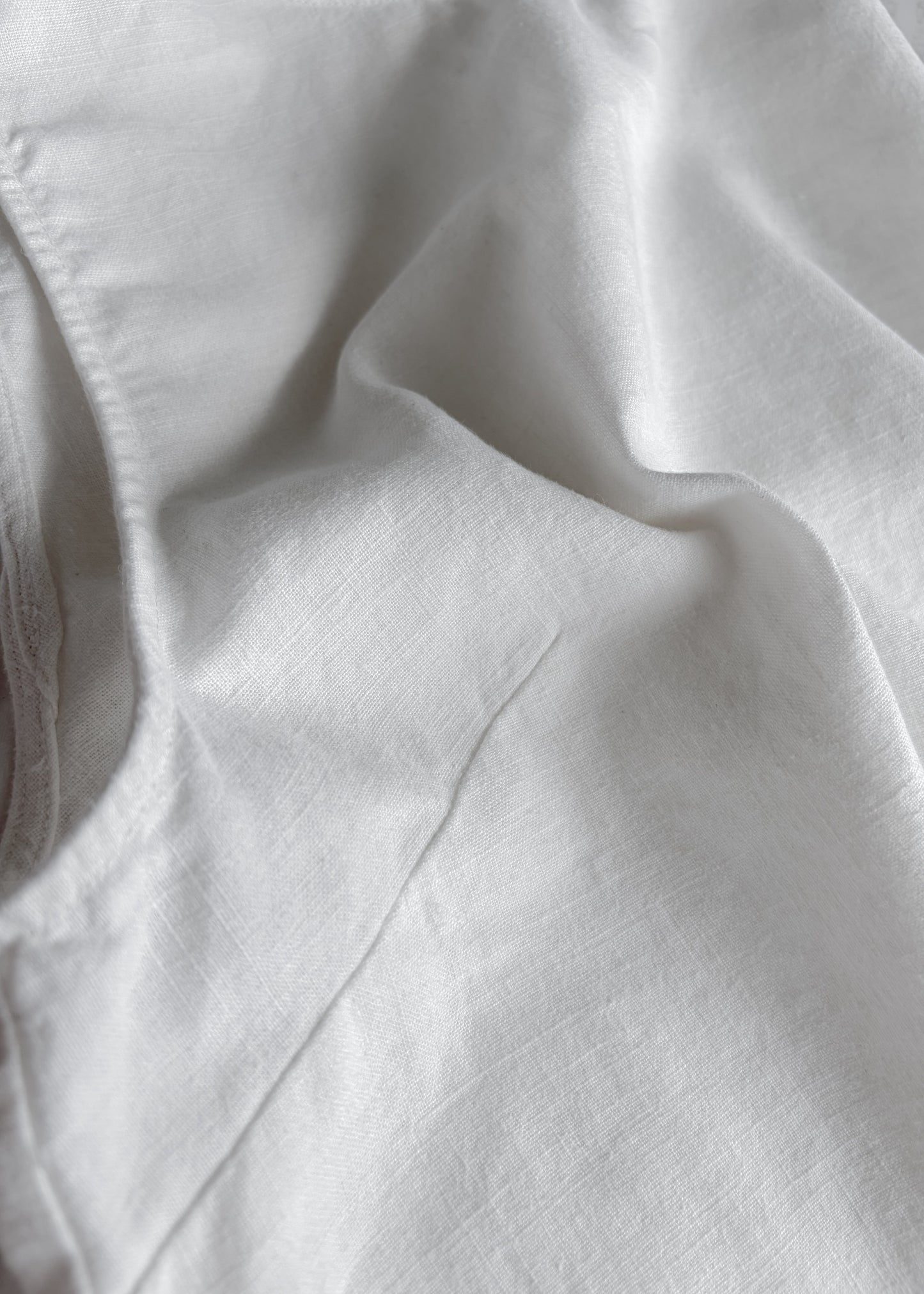 A close-up of white fabric with soft folds and a visible stitched edge on the left side.
